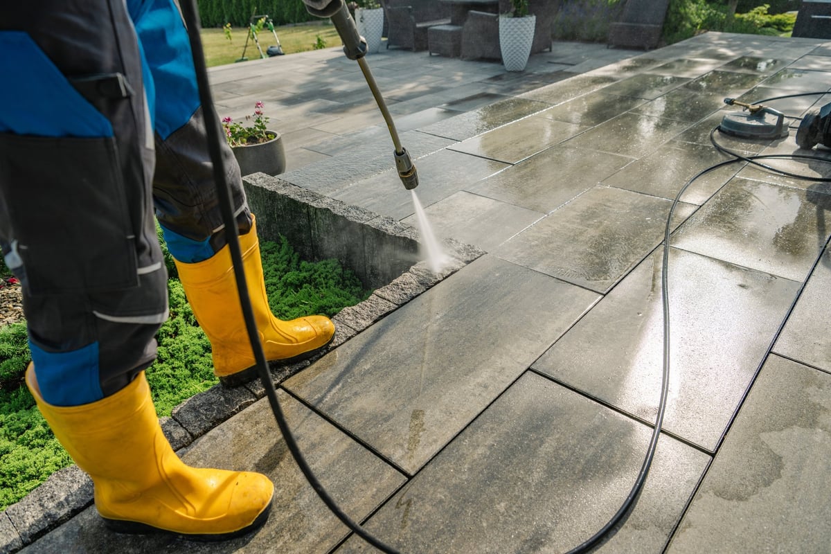 Worker in yellow boots pressure washing a stone patio surrounded by residential greenery